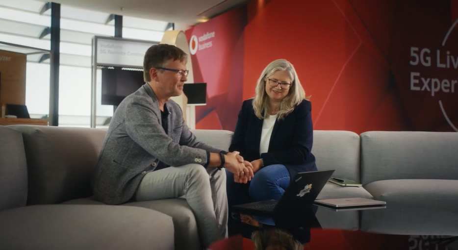 A man and woman seated on a couch in a professional setting. They smile while watching something on a laptop. Behind them is a red wall with a 