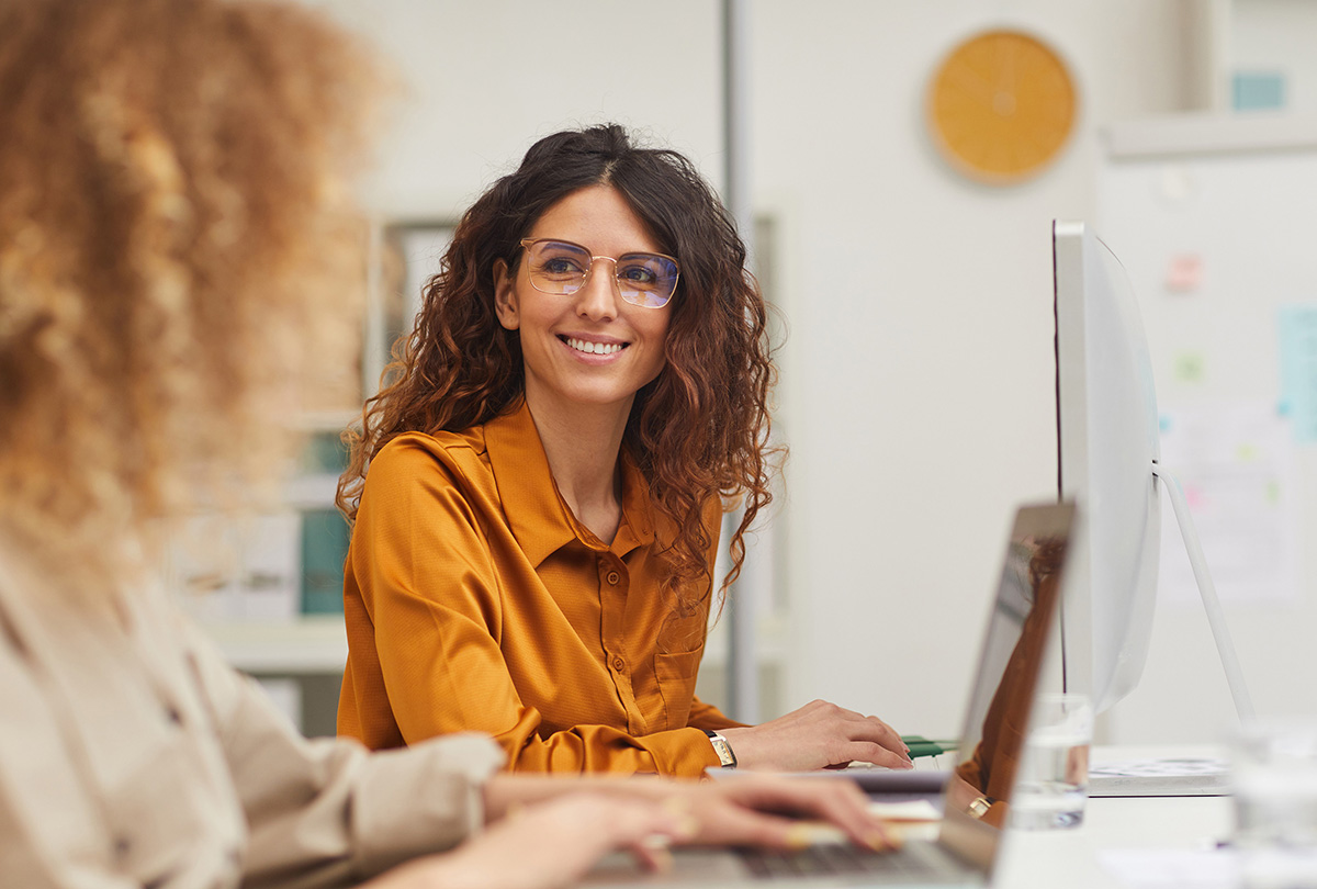 A smiling woman in an orange blouse and glasses sits at her desk in an office, with a computer monitor and the back of a colleague in the foreground.